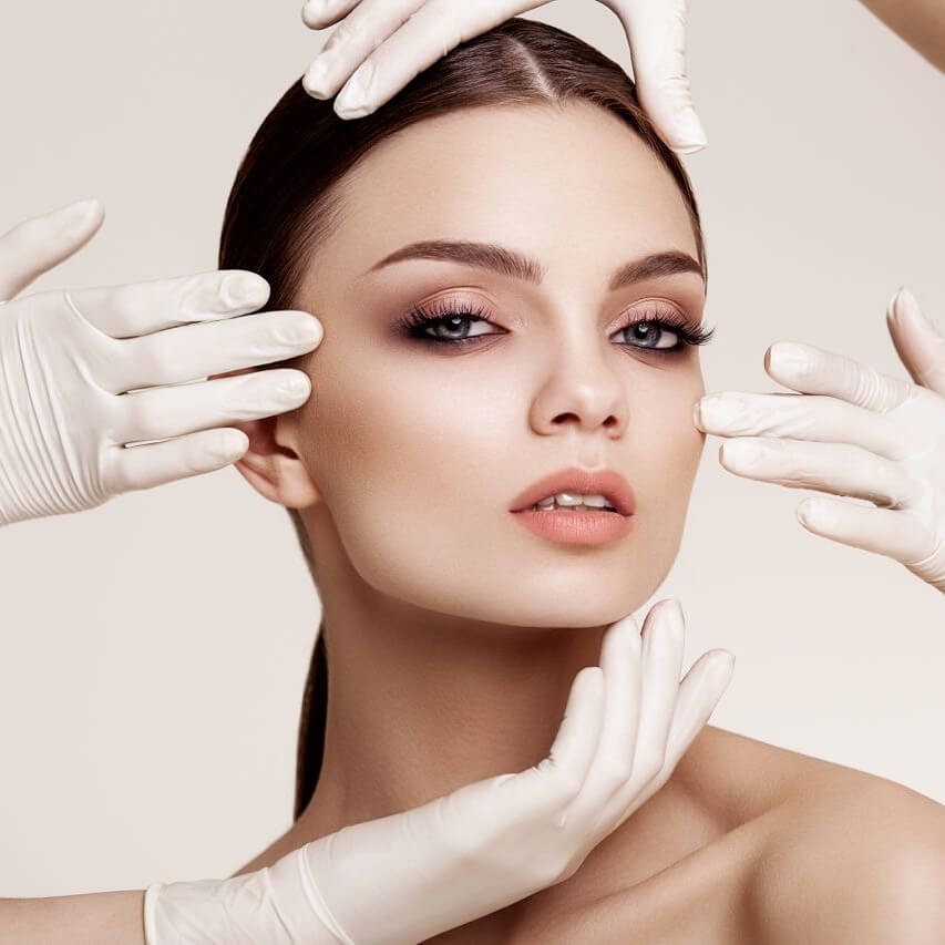Young brunette woman with wrinkle-free skin looks at the camera while four hands in white gloves touch various parts of her face and head